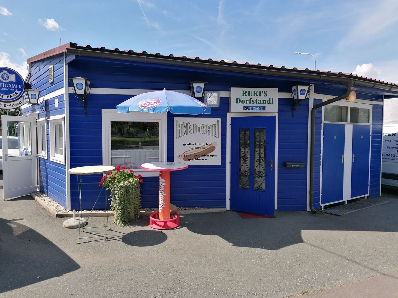 Ein blaues Gebäude mit einem Schild, das 'Ruki's Dorfstand' und eine blaue Tür zeigt. Draußen stehen zwei Tische mit einem Regenschirm und einem Topf. Das Gebäude hat ein rotes Dach und weiße Verkleidung.