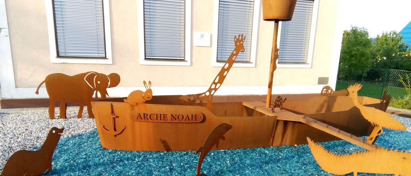 Eine Noahs-Arche-Skulptur ist vor einem Gebäude mit drei Fenstern ausgestellt und zeigt Tiere wie Elefant, Giraffe und Schnecke. Die Arche ist von blauem Kies umgeben.