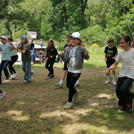 Eine Gruppe von Kindern spielt draußen auf einer Grasfläche, einige laufen im Kreis, andere gehen, mit Bäumen und einem Tisch im Hintergrund.