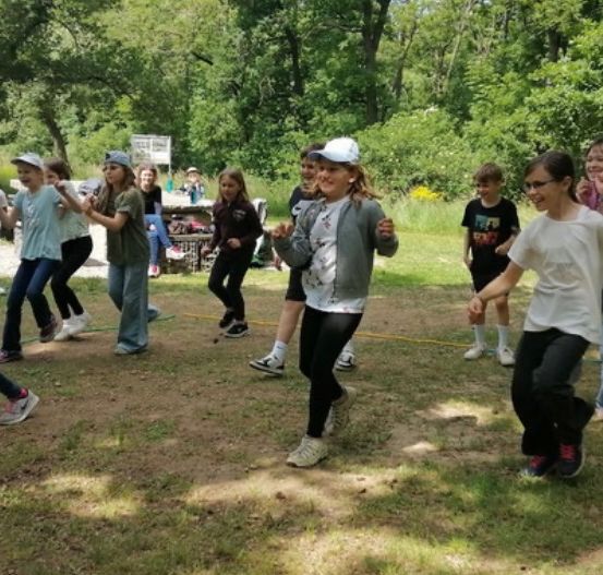 Eine Gruppe von Kindern spielt draußen auf einer Grasfläche, einige laufen im Kreis, andere gehen, mit Bäumen und einem Tisch im Hintergrund.