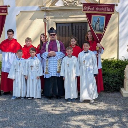 Ein Priester steht mit Messdienern und Messdienerinnen, die Banner halten, vor einer Kirche mit verzierten Säulen und einem Eingang.