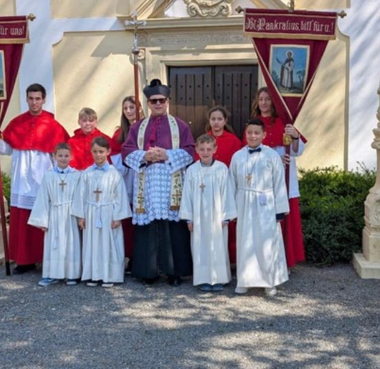 Ein Priester steht mit Messdienern und Messdienerinnen, die Banner halten, vor einer Kirche mit verzierten Säulen und einem Eingang.