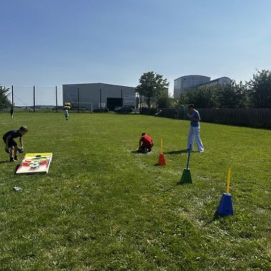 Eine Gruppe von Menschen spielt auf einem Rasenfeld mit einem Cornhole-Brett und Kegeln. Eine Person beugt sich hinunter, um etwas aufzuheben.