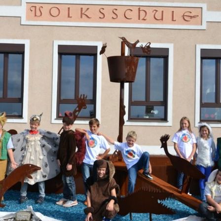 Mehrere Kinder in Kostümen stehen vor einem Gebäude mit dem Namen 'Volksschule'. Sie befinden sich neben einer Metallstruktur mit einem Vogel obenauf und einem blauen Teppich auf dem Boden. Einige tragen Tierkostüme.