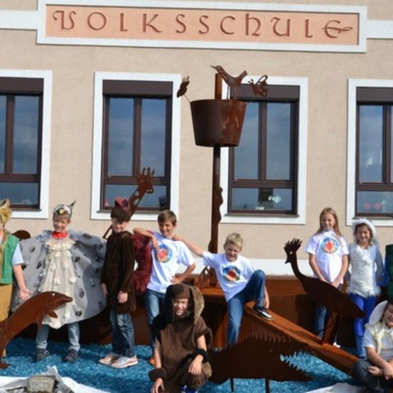 Mehrere Kinder in Kostümen stehen vor einem Gebäude mit dem Namen 'Volksschule'. Sie befinden sich neben einer Metallstruktur mit einem Vogel obenauf und einem blauen Teppich auf dem Boden. Einige tragen Tierkostüme.