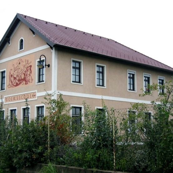 Ein beigefarbenes Gebäude mit einem roten Dach zeigt eine Wandmalerei von tanzenden Menschen. Der Text 'Volksschule' steht über dem Eingang. Es gibt mehrere Fenster und eine Lampe an der Wand.