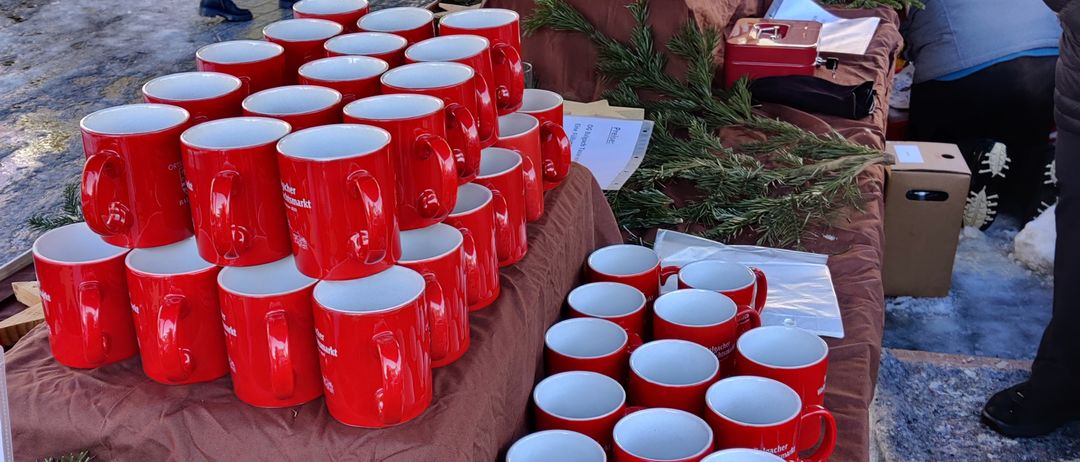 Rote Tassen sind in Stapeln auf braunen Tischdecken ausgestellt, mit Grünpflanzen und Tannenzweigen. Ein Kinderwagen und Menschen sind im Hintergrund.