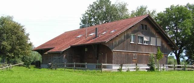 Ein altes Holzhaus mit rotem Dach und mehreren Fenstern ist von einem Holzzaun umgeben in einem grünen Feld mit Bäumen im Hintergrund.