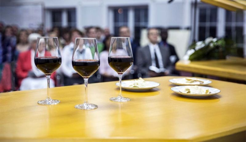 Drei mit rotem Wein gefüllte Weingläser stehen auf einem Tisch vor einem sitzenden Publikum.