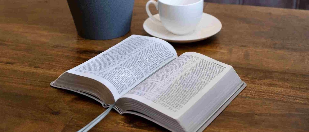 Ein offenes Buch liegt auf einem Holztisch mit einer Tasse und Untertasse neben einem Topf.