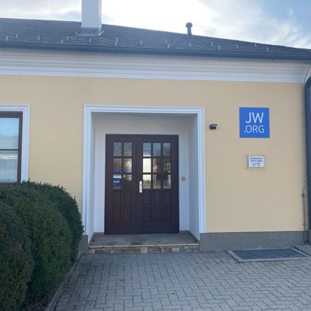Die Vorderansicht eines Gebäudes mit einem blauen Schild, auf dem JW.ORG steht. Der Eingang hat eine Doppeltür mit Glasscheiben.