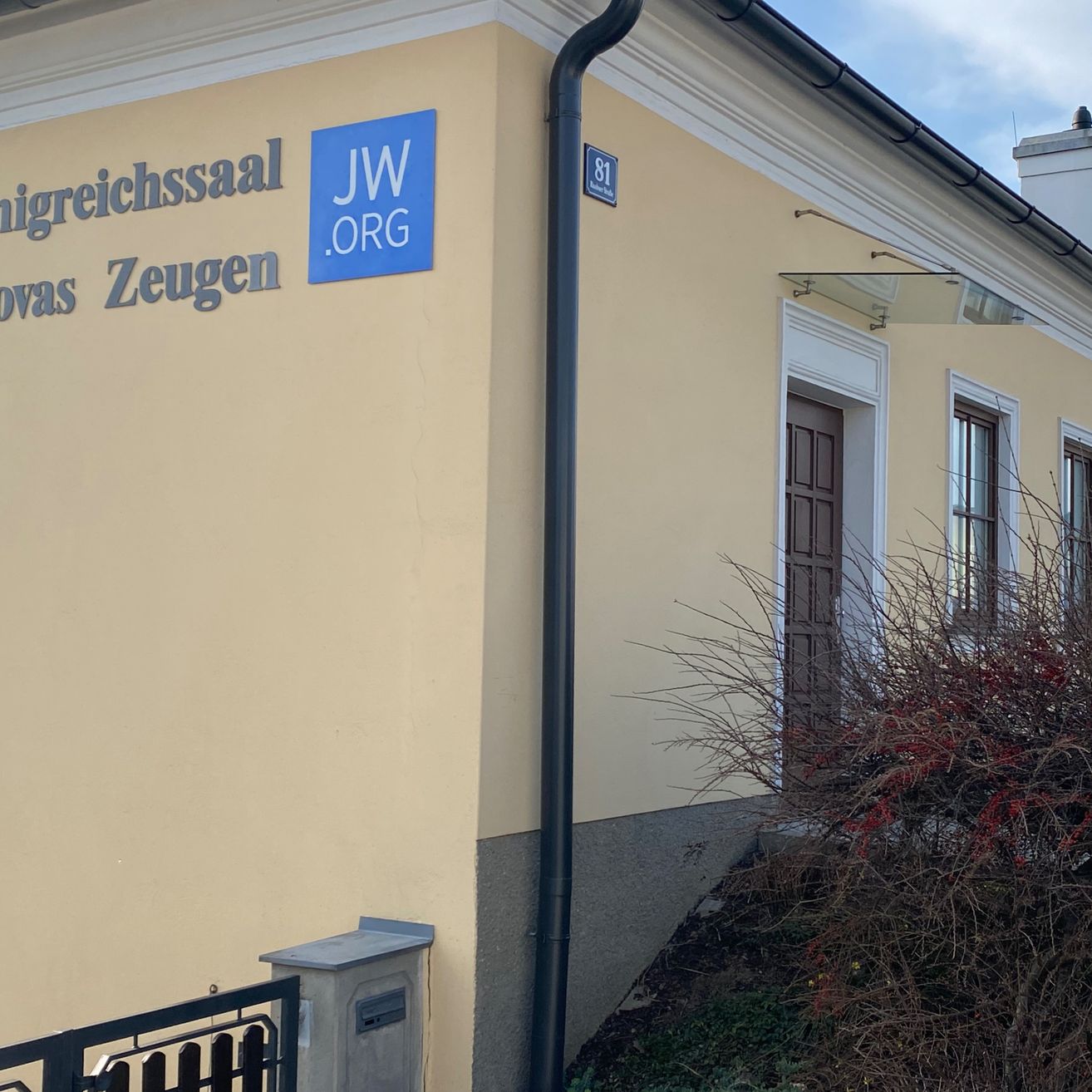 Die Seite eines Gebäudes mit einem blauen Schild, auf dem 'JW.ORG' und die Nummer '81' stehen. Unter dem Schild sind die Worte 'Nigreichsaal' und 'ovas Zeugen' zu sehen. Es gibt eine braune Tür und zwei Fenster.