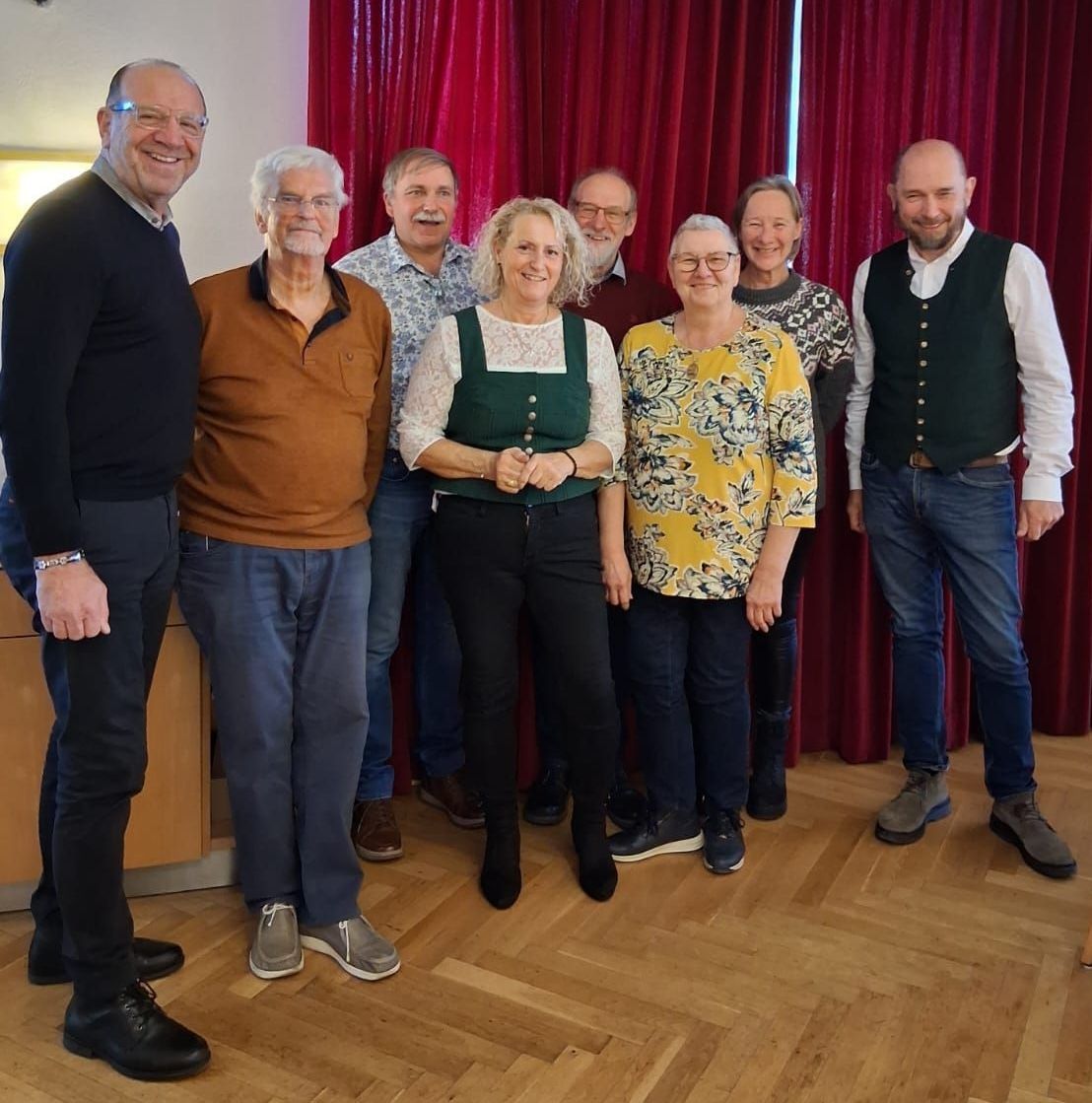 Eine Gruppe von sieben Personen steht lächelnd zusammen auf einem Holzboden. Sie sind lässig gekleidet, zwei Frauen tragen Schürzen. Ein roter Vorhang ist hinter ihnen.