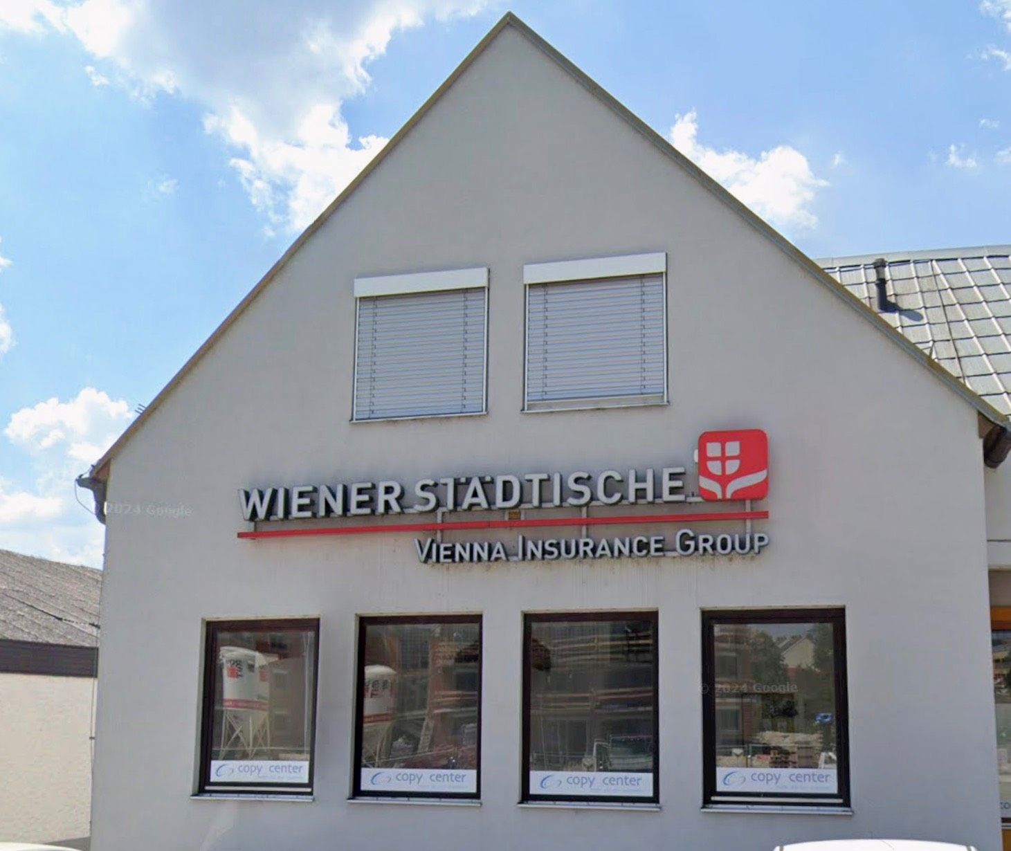 Die Vorderseite eines Gebäudes mit einem Schild, das Wiener Stadtische Vienna Insurance Group liest. Das Gebäude hat vier Fenster und zwei Rolläden über den Fenstern. Die Fenster sind geschlossen und die Rolläden sind geschlossen.