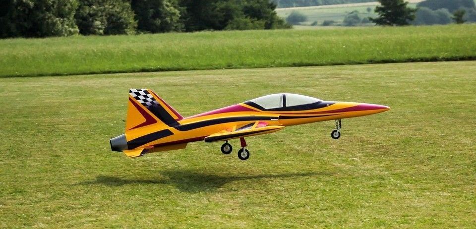 Ein farbenfrohes ferngesteuertes Flugzeug fliegt auf einer weiten, offenen Wiese mit Bäumen im Hintergrund.