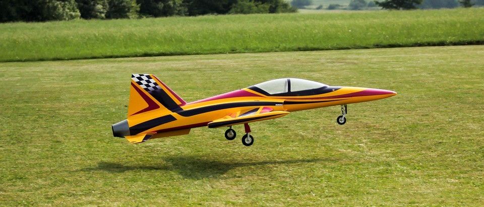 Ein gelbes und rotes Modellflugzeug mit einem karierten Schwanz fliegt über ein Grasfeld mit Bäumen im Hintergrund.