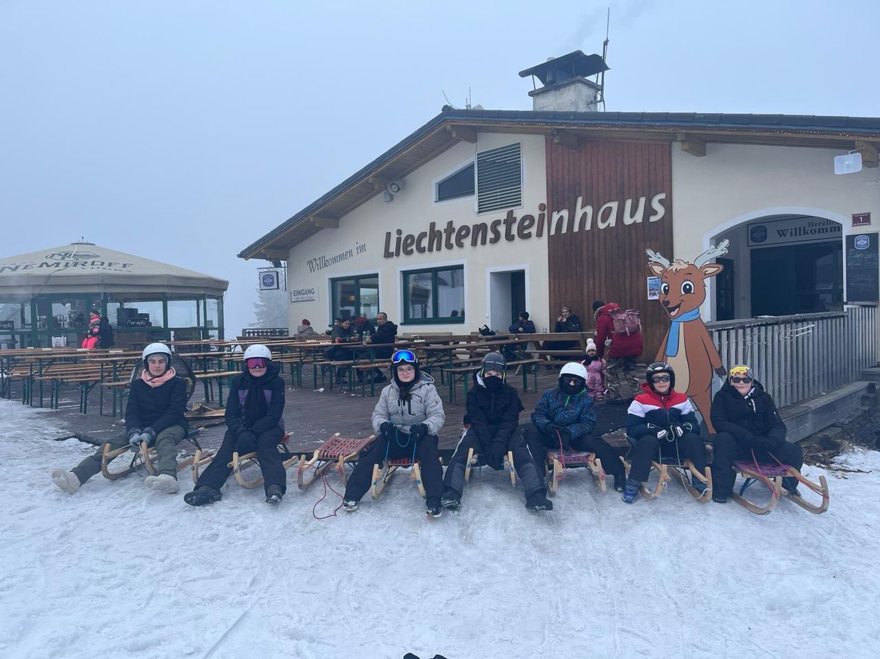 Eine Gruppe von Menschen sitzt auf Schlitten vor einem Gebäude namens Liechtensteinhaus an einem verschneiten Tag. Sie sind alle in Winterkleidung gekleidet und tragen Helme. Es gibt ein Rehkitz-Maskottchen am Gebäude.