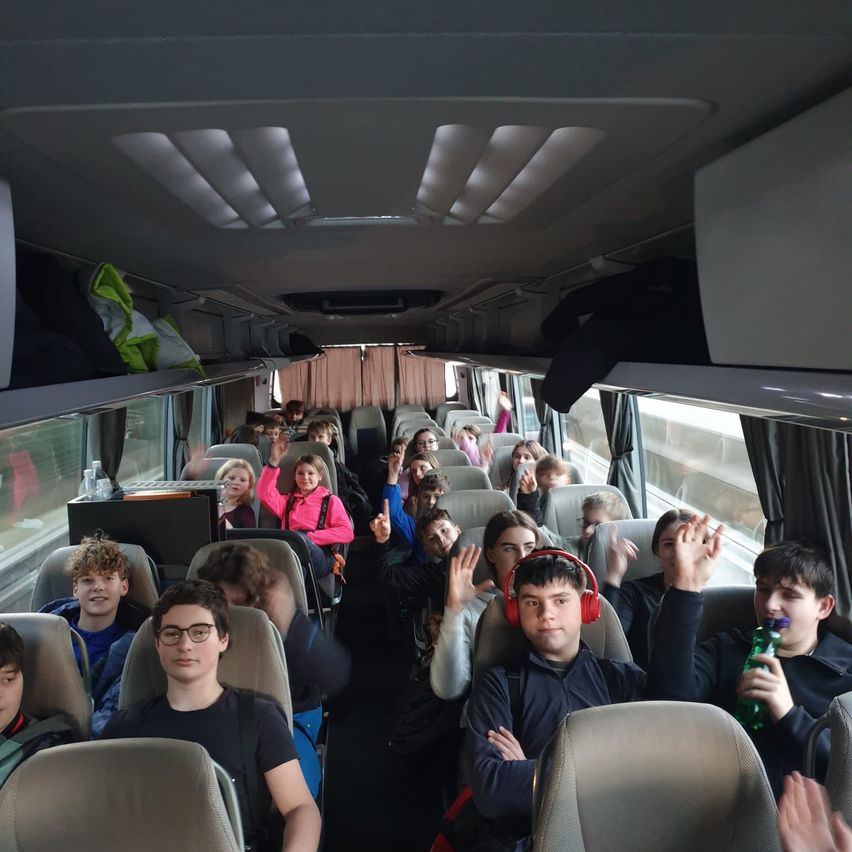 Ein Schulbus voller Schüler auf einer Reise, einige mit Kopfhörern, andere mit erhobenen Händen, einige mit Rucksäcken und einige schauen aus dem Fenster.