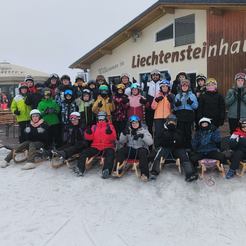 Eine Gruppe von Menschen sitzt auf Schlitten vor einem Gebäude namens Liechtensteinhaus. Sie sind alle in Winterkleidung und Helmen gekleidet. Einige posieren für ein Foto.