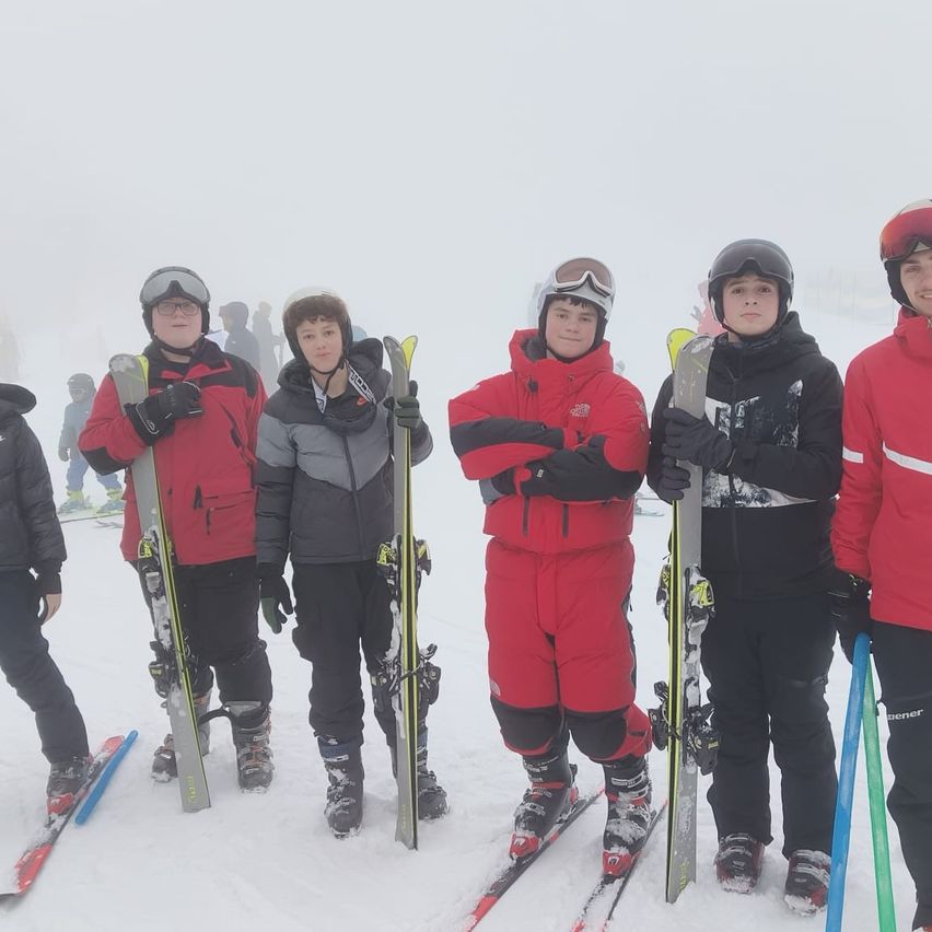 Fünf junge Skifahrer stehen im Schnee, tragen Winterkleidung und Helme. Sie halten Skistöcke und Skier in der Hand. Der Hintergrund ist neblig.