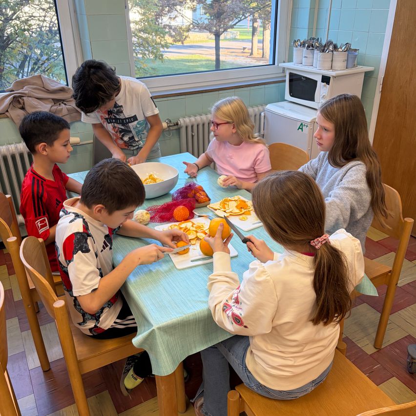 Eine Gruppe von Kindern sitzt um einen Tisch herum und schält Orangen. Ein Junge hält ein Messer und schneidet eine Orange. Der Tisch hat eine blaue Tischdecke und ist mit einer Schüssel voller Orangen bedeckt. Hinter ihnen befindet sich ein Fenster mit Blick auf Bäume.
