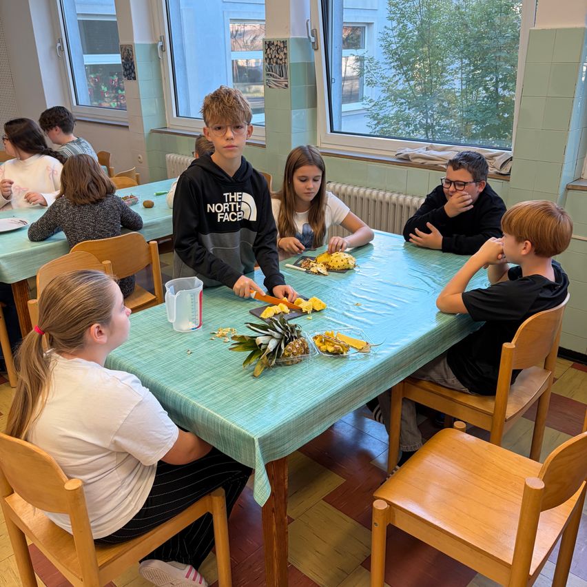 Eine Gruppe von Kindern sitzt um einen Tisch mit Ananas und Messern. Ein Kind schneidet die Ananas.