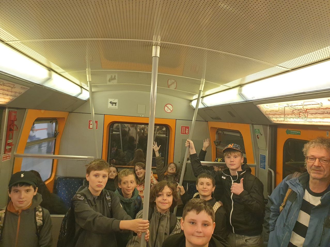 A group of children and young people are riding a train. They are posing for a photo, some with hands raised, others with thumbs up. The train is filled with passengers.