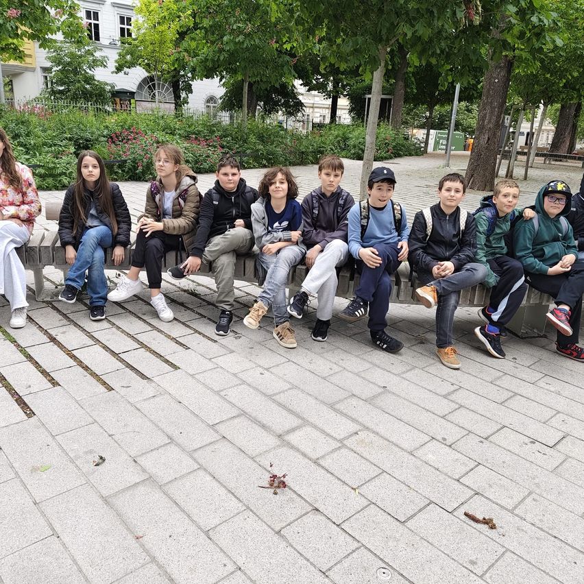 A group of young people are sitting on a concrete bench in a park. They are all dressed in winter clothing. There are trees and plants in the background.