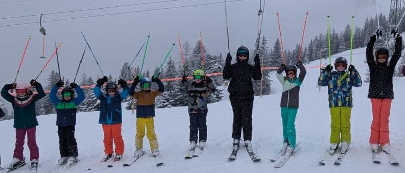 Eine Gruppe von Kindern und Erwachsenen in Winterkleidung posiert für ein Foto auf einer verschneiten Piste mit Skiliften im Hintergrund.