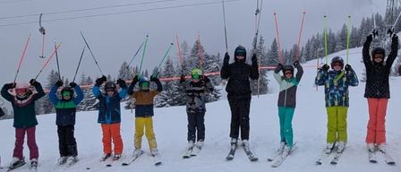 Eine Gruppe von Kindern und Erwachsenen in Winterkleidung posiert für ein Foto auf einer verschneiten Piste mit Skiliften im Hintergrund.