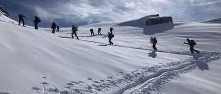 Eine Gruppe von Skifahrern in Ausrüstung erklimmt einen verschneiten Berg. Sie verwenden Skistöcke, um sich im Schnee zurechtzufinden. Der Himmel ist bewölkt mit einer hellen Sonne im Hintergrund.