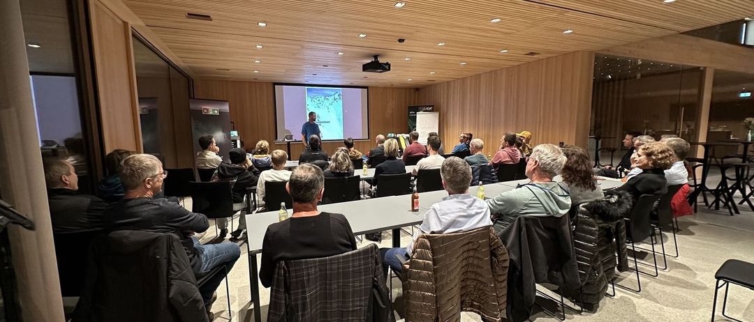 Eine Gruppe von Menschen nimmt in einem Raum mit Holzwänden an einer Präsentation teil. Der Präsentator steht vor einer Leinwand und die Teilnehmer sitzen in Stühlen.