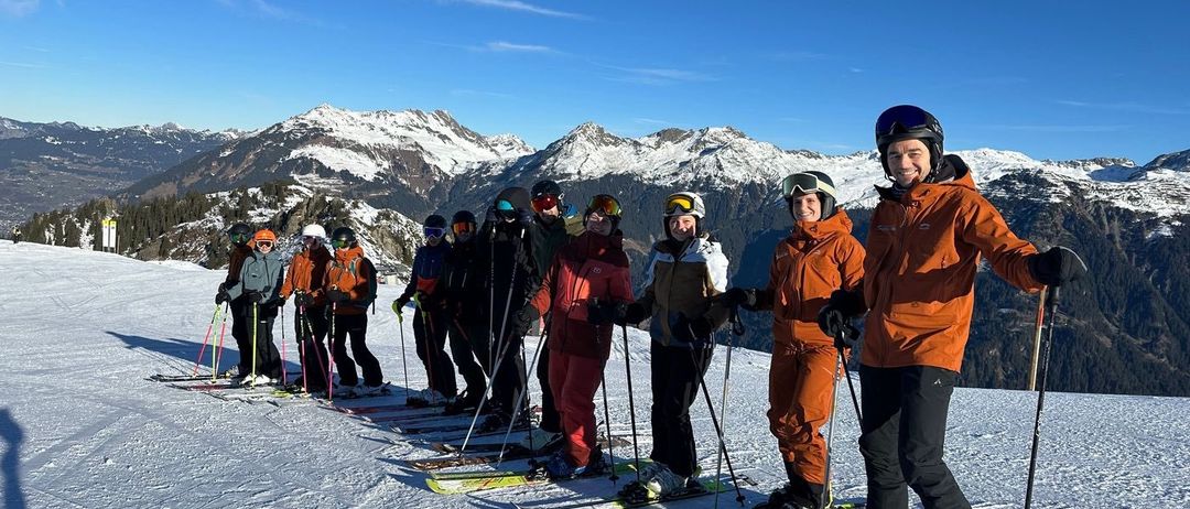Eine Gruppe von Skifahrern in orangefarbenen Jacken posiert auf einem verschneiten Berg. Sie halten Skistöcke und tragen Helme. Im Hintergrund sind schneebedeckte Berge unter einem klaren blauen Himmel zu sehen.