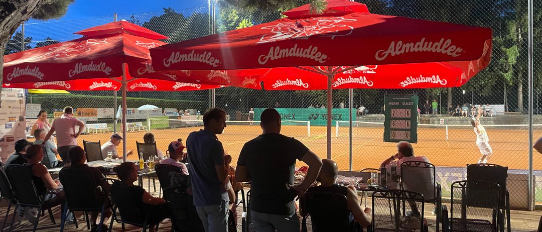 Ein Tennisplatz im Freien mit Menschen unter Sonnenschirmen. Eine Anzeigetafel ist an der Umzäunung zu sehen. Einige Personen stehen, während andere sitzen. Bäume und ein Zaun umgeben den Platz.