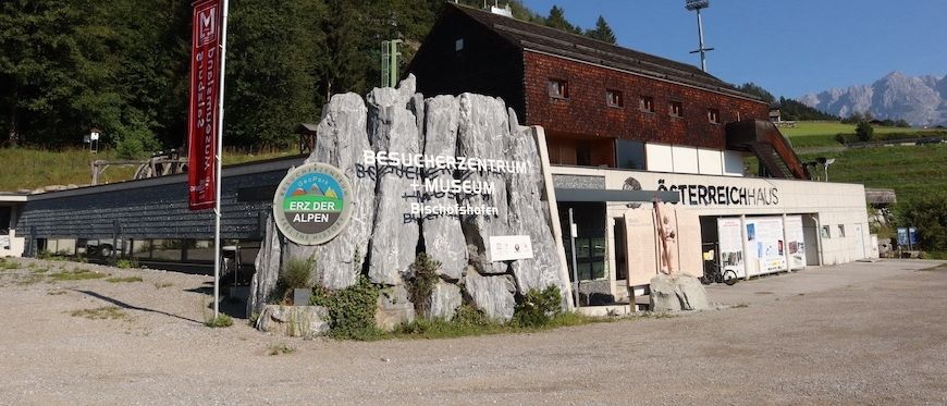 Der Eingang zu einem Besucherzentrum und Museum ist zu sehen, mit einer großen Steinstruktur und einem roten Banner davor. Dahinter ist ein Gebäude mit einer Treppe und einem Schild mit der Aufschrift 'Sterreichhaus' zu erkennen.