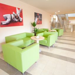 Ein Innenraum mit grünen Ledersofas, Tischen und einer Treppe. Ein roter Poster von Menschen in Kampfsportausrüstung ist an der Wand.