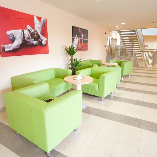 Ein Innenraum mit grünen Ledersofas, Tischen und einer Treppe. Ein roter Poster von Menschen in Kampfsportausrüstung ist an der Wand.
