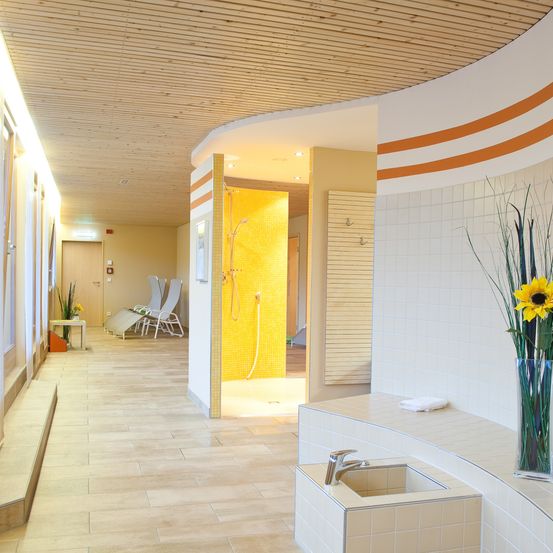 Ein modernes Spa mit gelben und weißen Fliesen, einer Dusche und einem Waschbecken. Es gibt Liegestühle und einen Tisch mit einer Pflanze. Die Decke ist aus Holz.