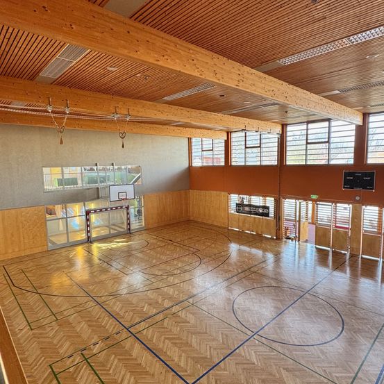 Ein leerer Indoor-Basketballplatz mit Holzboden und Balken, großen Fenstern und einem Basketballkorb.