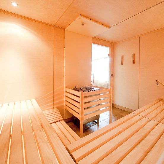 Ein leerer Saunaraum mit Holzbänken und einem kleinen Holzregal, beleuchtet von sanftem Licht.