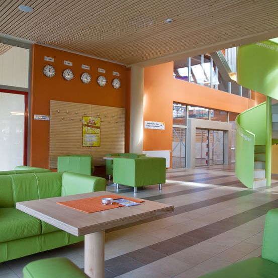 Ein Innenbereich mit grünen Sofas, einem Holztisch und einer Wendeltreppe. Die Wände sind orange und weiß gestrichen, und es hängen Uhren an der Wand.