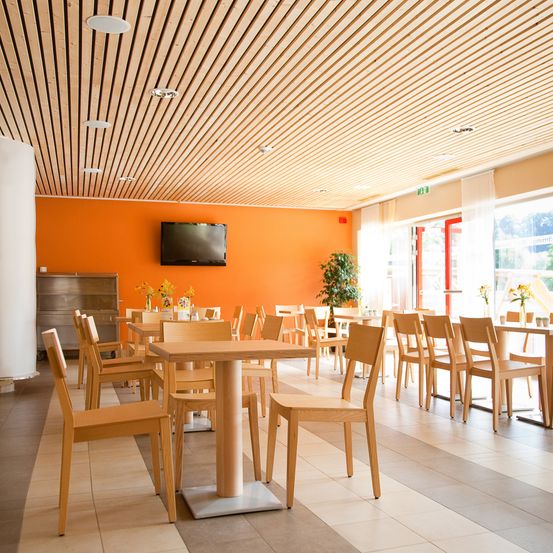 Ein leerer Essbereich mit Holztischen und -stühlen, einer orangefarbenen Wand, einem montierten Fernseher, einer Topfpflanze und einem weißen Pfeiler.