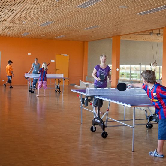 Mehrere Personen spielen Tischtennis in einem Raum mit orangefarbenen Wänden. Eine Frau serviert den Ball, während andere auf ihren Zug warten.