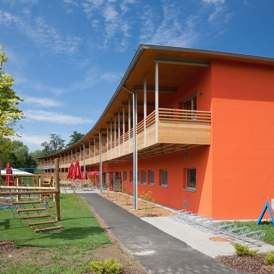 Ein orangefarbenes Gebäude mit kurvenförmiger Gestaltung, das hölzerne Balkone und Metallgeländer aufweist. Es ist von einem Rasen umgeben, der ein Spielplatz und ein Weg ist. Das Gebäude hat mehrere Fenster und einen kleinen Garten davor.