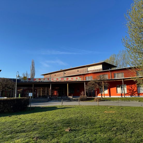 Das Sportcampus-Gebäude, in Orange gestrichen, steht prominent vor einem klaren blauen Himmel. Es verfügt über ein Schild und große Fenster, wobei Bäume und Büsche die Vorderseite einrahmen.