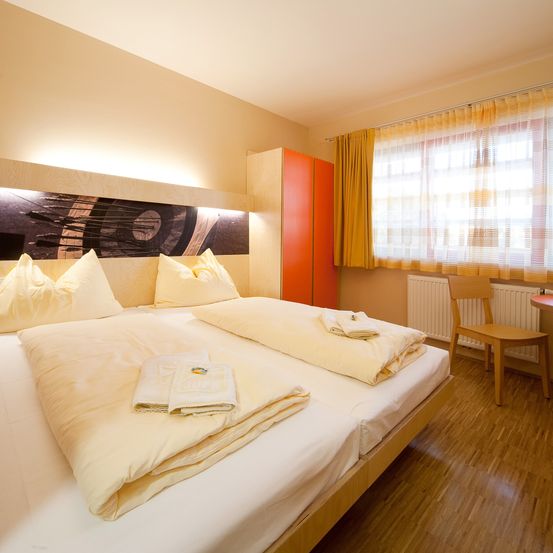 Ein Schlafzimmer mit zwei Betten, cremefarbener Bettwäsche und Kissen. Das Kopfteil des Bettes hat ein dunkles, künstlerisches Design. Ein Holzstuhl und ein Heizkörper befinden sich am Fenster.