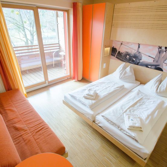 Ein Hotelzimmer mit zwei Betten, einer Couch und einem Balkon. Das Zimmer hat ein modernes Design mit orangefarbenen Akzenten.