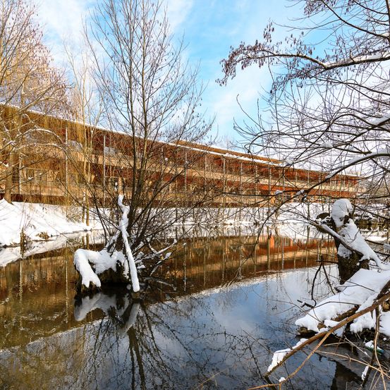 Ein Gebäude mit mehreren Stockwerken und Balkonen befindet sich neben einem Fluss. Das Gebäude und die Bäume sind schneebedeckt. Der Fluss spiegelt das Gebäude und die Bäume wider.