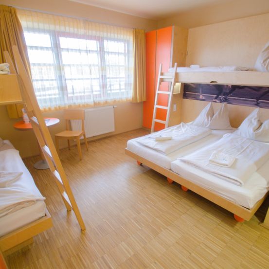 Ein Schlafsaal mit Etagenbetten, einer Leiter und einem Fenster mit Vorhängen. Ein Stuhl und ein kleiner Tisch stehen neben dem Fenster. Der Raum hat Holzböden.