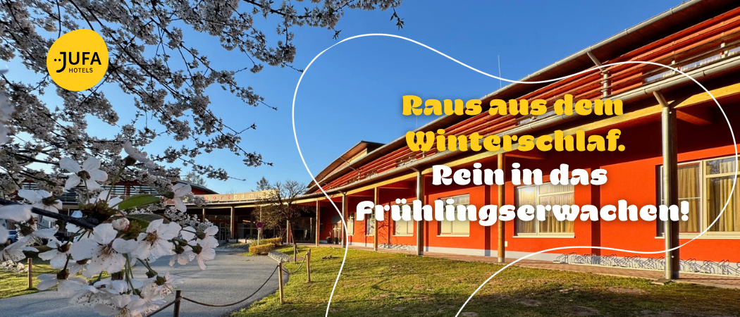 Ein Gebäude im Freien mit Rasenfläche davor und einem blühenden Baum. Ein Text-Overlay sagt 'Raus aus dem Winterschlaf. Rein in das Frühlingserwachen.'